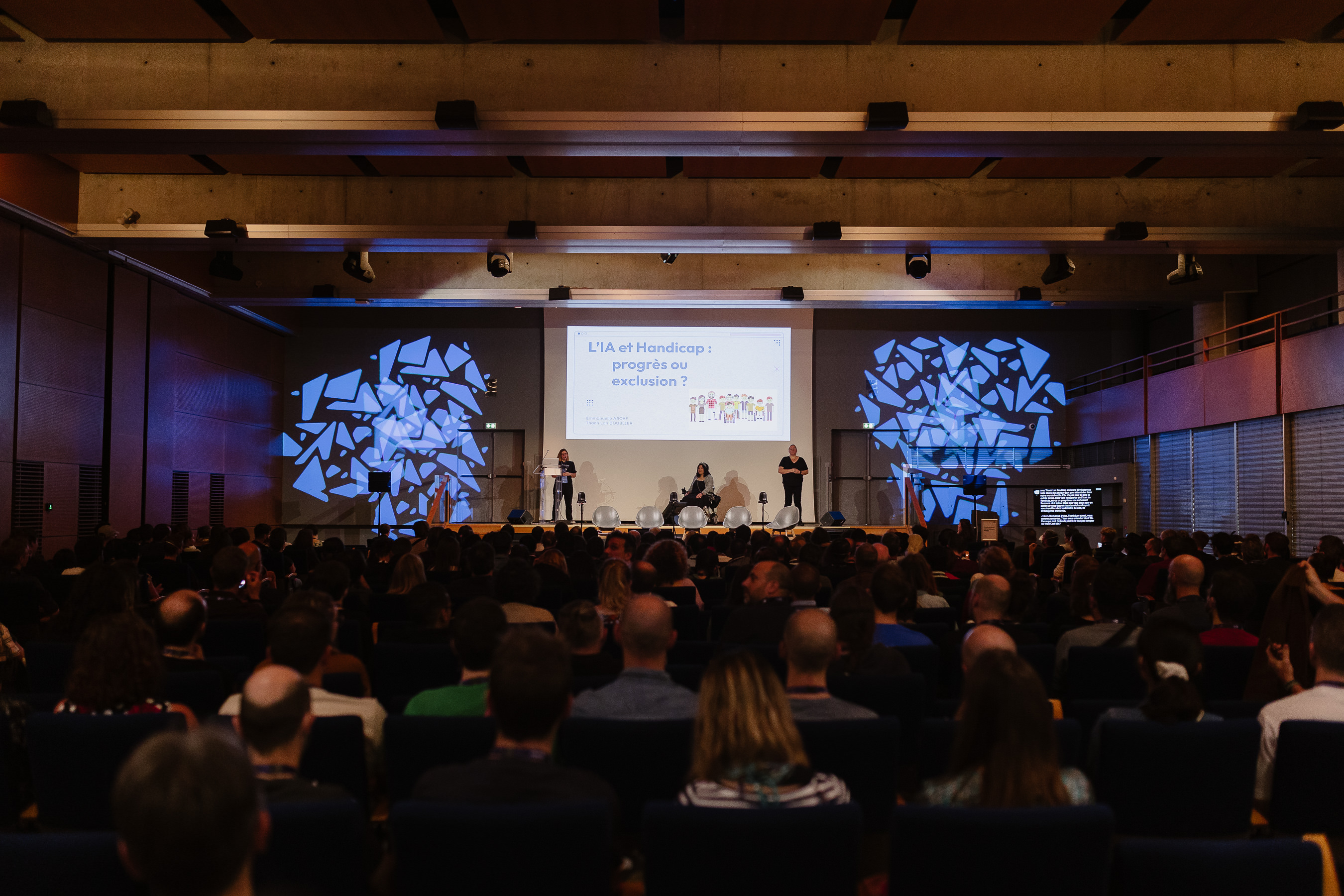On voit le public de dos et au loin la scène avec Emmanuelle, Thanh Lan et une interprète en LSF. Derrière elles est projetée la présentation L'IA et Handicap : progrès ou exclusion. Sur la droite au fond, on voit un écran de vélotypie.