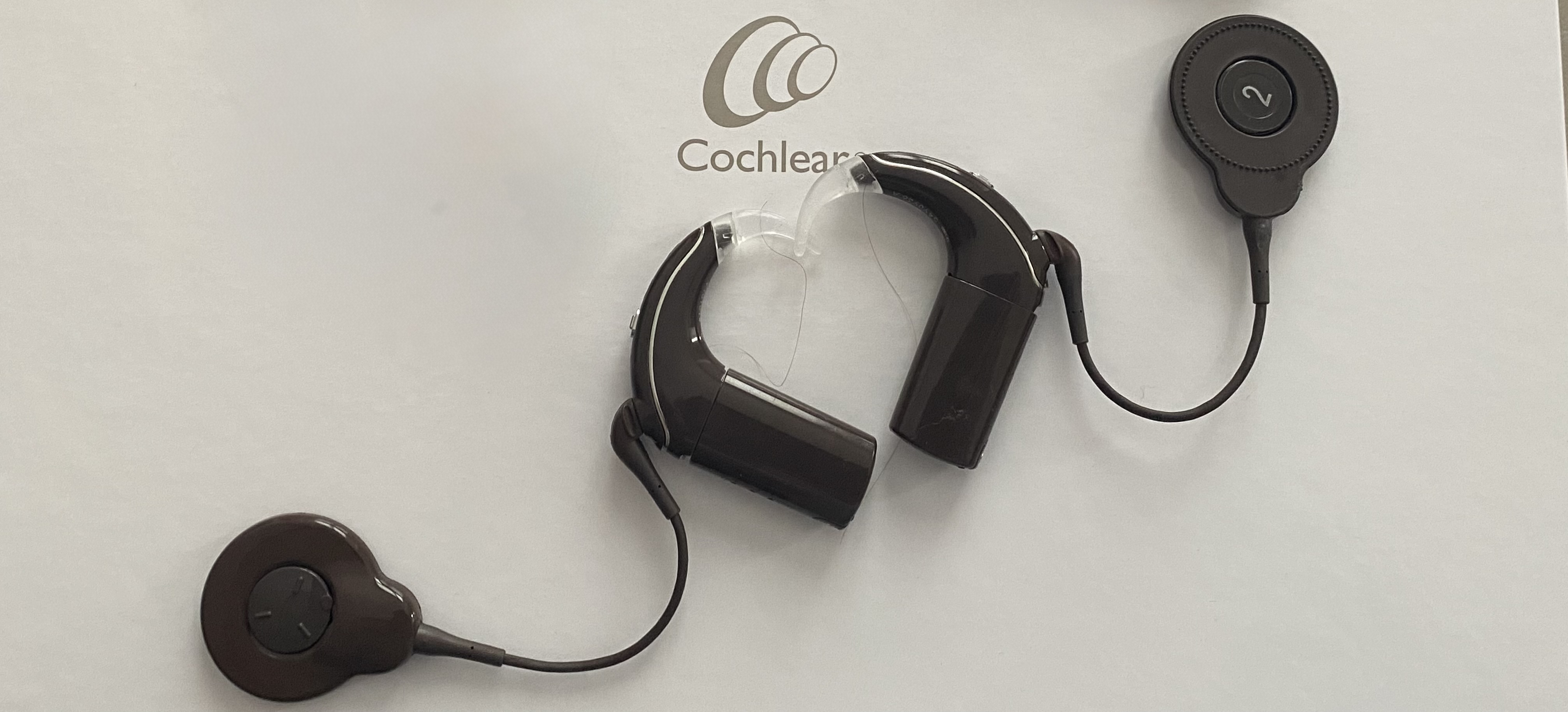  Une nouvelle version de mes deux implants cochléaires de couleur marron qui sont côte à côte et forment un coeur. En arrière plan, la couleur blanche et on voit la marque Cochlear.