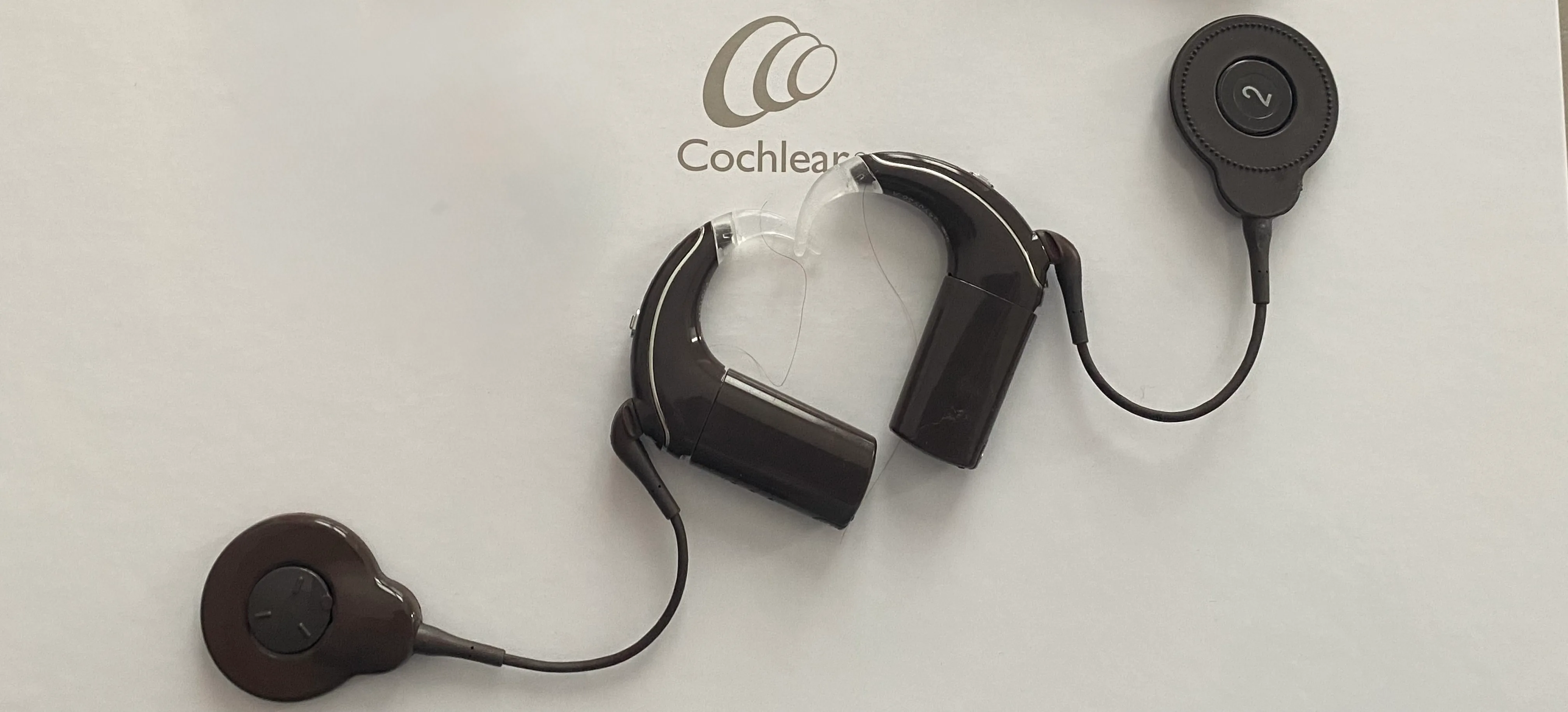  Une nouvelle version de mes deux implants cochléaires de couleur marron qui sont côte à côte et forment un coeur. En arrière plan, la couleur blanche et on voit la marque Cochlear.