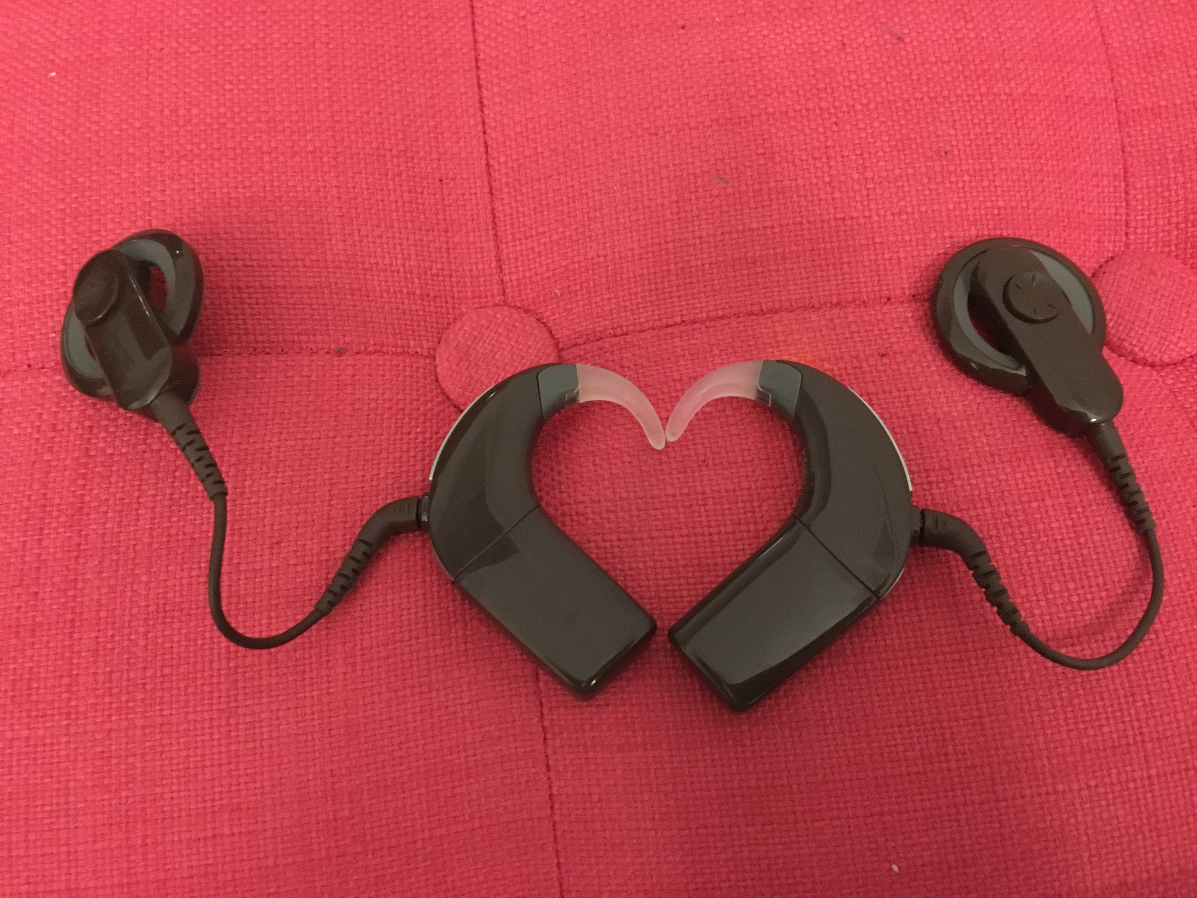 Mes deux implants cochléaires de couleur marron sont côte à côte et forment un coeur. En arrière plan, la couleur rose.