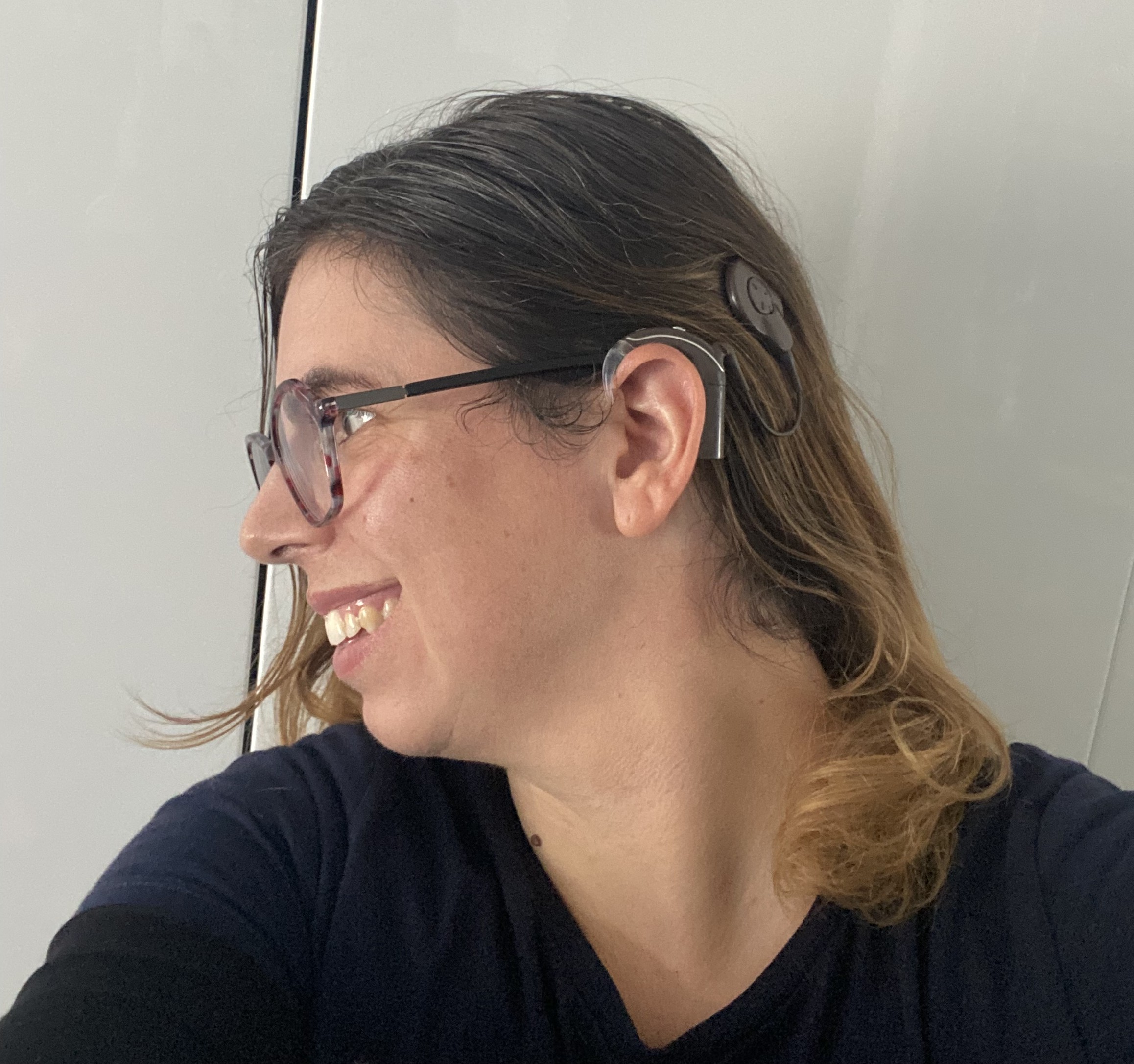 Je suis de profil. Mes cheveux châtains sont longs jusqu’aux épaules. Je suis souriante et j’ai des lunettes. On voit mon implant cochléaire dont le contour est posé sur l’oreille avec un fil qui relie jusqu’à l’antenne collé sur les cheveux un peu en hauteur.