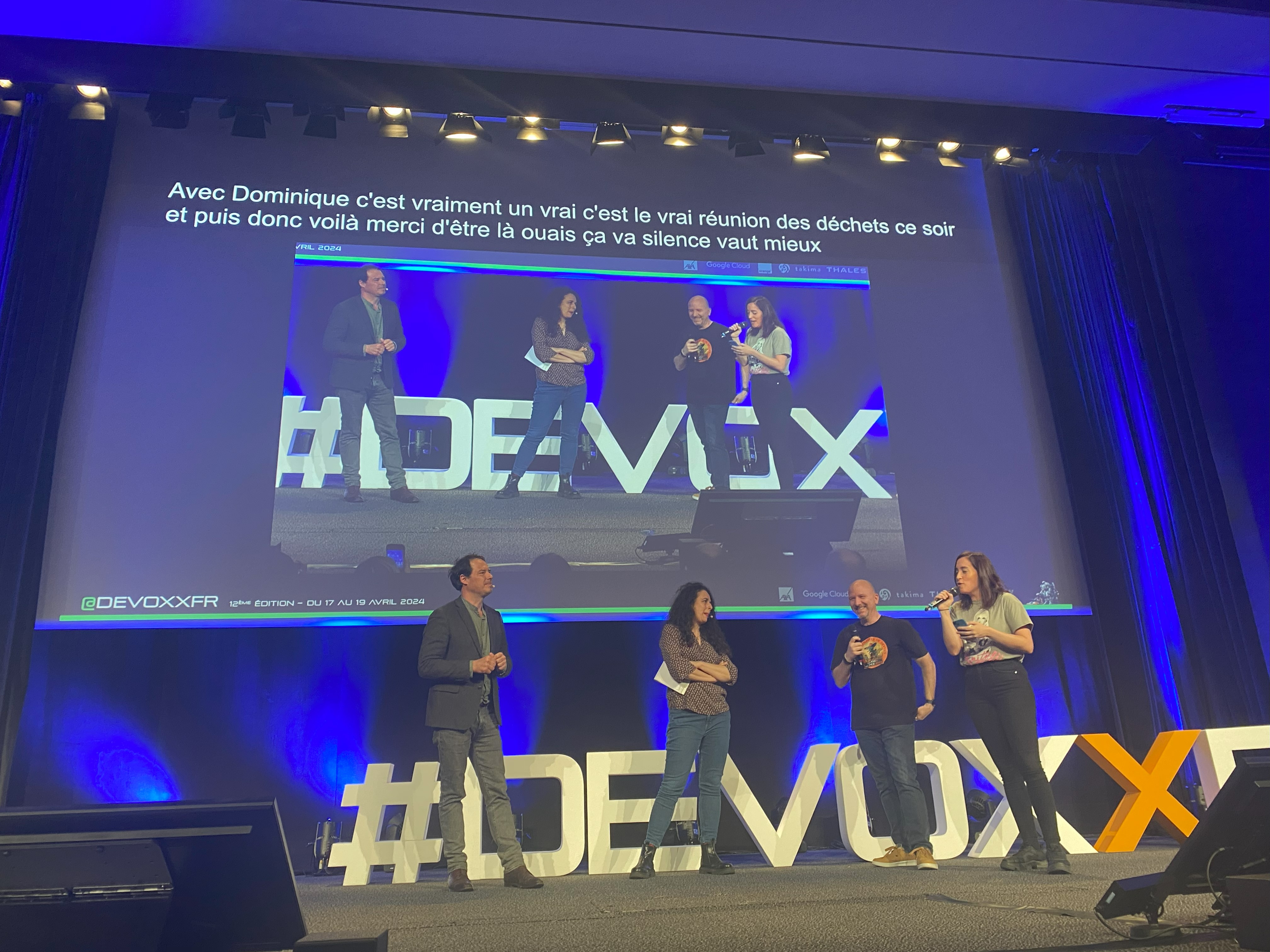 On voit 4 personnes dont 2 hommes et 2 femmes sur scène avec les lettres énormes #DEVOXXFR derrières elles. En dessus d'elles, l'écran où est projeté en live ces personnes. En dessus de l'écran, le bandeau noir des sous-titres automatiques transcrits après l'image.